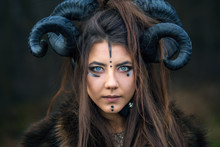 Outdoor Portrait Of Beautiful Scandinavian Viking Woman Warrior With Blue Eyes Wearing Ram Horns Looking At Camera. Female Hunter With Specific Makeup Wearing Fur Collar