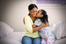 Smiling Mid-adult Woman Being Kissed On The Cheek By Her Daughter While Sitting With Their Arms Around Each Other On A Bed.