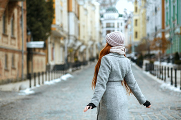 Wall Mural - Pretty red head woman wearing winter cap and coat posing at the city. View from the back. Empty space