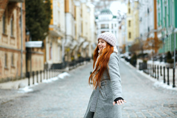 Wall Mural - Pretty red haired model wearing stylish winter outfit walking in city, spinning around. Empty space