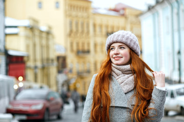 Wall Mural - Street portrait of joyful redhead girl with long hair wearing warm winter apparel posing at the street. Empty space