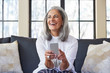 © Trinette Reed/Stocksy - Mature woman with grey hair texting on cell phone in living room at home