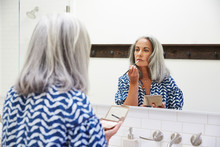 Senior Woman With Grey Hair Putting On Makeup In Bathroom Mirror