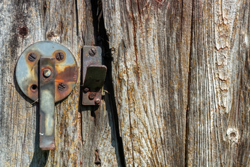 A rusty round swing latch is mounted on the door of a weathered brown barn siding with 4 rusty screws. The vertical latch is hanging down. The opposite catch is mounted with 2 rusty screws. 