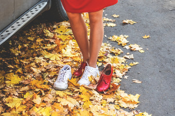 Young woman putting on white sports shoes for active walking in park. Legs of single girl in run snickers on the road among falling yellow maple leaves. Red woman boots wiht high heels. Autumn.