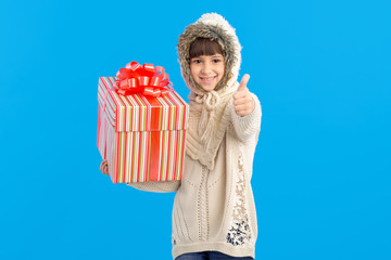 Wall Mural - Little girl with a big gift box in her hands showing thumb up on a blue background. Like. Christmas holiday gifts.