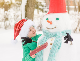 Wall Mural - portrait of smiling boy in red christmas hat holds gift box and hugs snowman