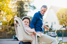 Mother And Daughter Riding Bicycle, Baby Wearing Helmet Sitting In Children's Seat