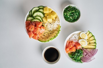 Raw Organic Poke Bowl with with rice, avocado, salmon, mango, cucumbers, chuka salad, quail eggs, sweet onions and soy sauce two plates close-up on white table background. Top view