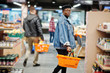 Stylish casual african american man at jeans jacket and black beret holding two baskets, walking and shopping at supermarket.