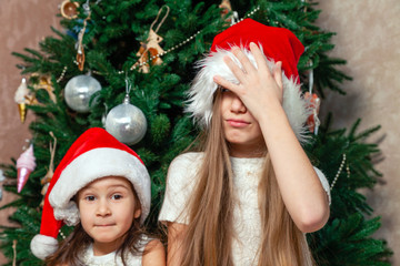 Two girlfriends posing and fooling around the New Year tree near the fireplace in a luxurious country mansion in funny red hats. The concept of strong friendship and devotion since childhood