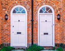 Two White Wood Residential Front Doors, With A Drain Pipe Down The Middle. The Walls Are Red Brick There Are Lunette Arches Over The Doors And Metal Lion Door Knockers