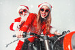 © proimagecontent - Snata Claus waering sunglasses with gifts bag sitting on the bike after smiling sexy girl in red hat, fun Christmas holidays concept