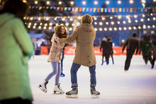 Young Couple In Love Caucasian Man With Blond Hair With Long Hair And Beard And Beautiful Woman Have Fun, Active Date Skating On Ice Scene In Town Square In Winter On Christmas Eve