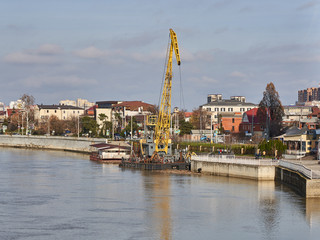 A yellow crane on a barge is standing on the Kuban river pier near the picturesque street of the Kuban Embankment in Krasnodar. The blue sky is like a watercolor painting.