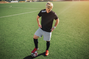 Wall Mural - Young blonde player stand on lawn and look down. He poses. Guy hold hands on hips. He is on lawn alone. Sun is shining outside.