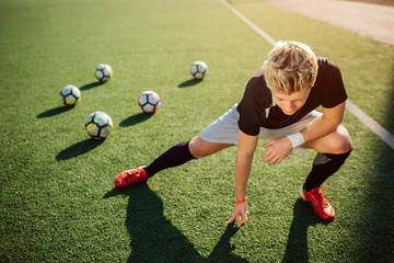 Wall Mural - Young player stretch on green lawn. He does exercises for legs. Guy look down. Four balls are behind him.