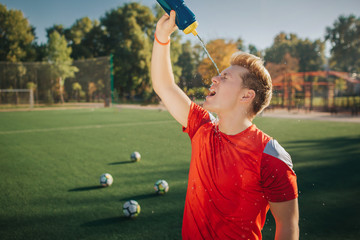 Wall Mural - Tired football player pouring water on face. He shrinks. Young man stand on lawn. Four balls are behind him.