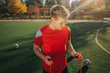 Wall Mural - Blonde young man holding jump rope iin hands. He looks at it. Guy stand on lawn outside. Sun is shining. Balls are all around young man.