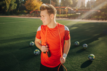 Wall Mural - Well-built athlete looks to left. He hold jump rope around neck. Guy stand on green lawn outside. Five balls lying behind. Sunny weather is outside.