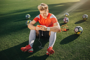 Wall Mural - Young player sit alone on green lawn. He has rest. Guy hold phone in hands with headphones. Six balls are behind him.