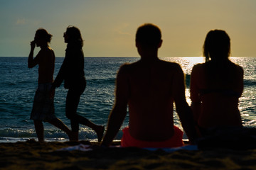 Silhouettes of a couple, man and woman are sitting on a sandy beach and looking at the sunset on the sea on a vacation. 