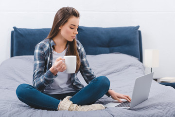 beautiful young woman holding mug with hot beverage and using laptop on bed