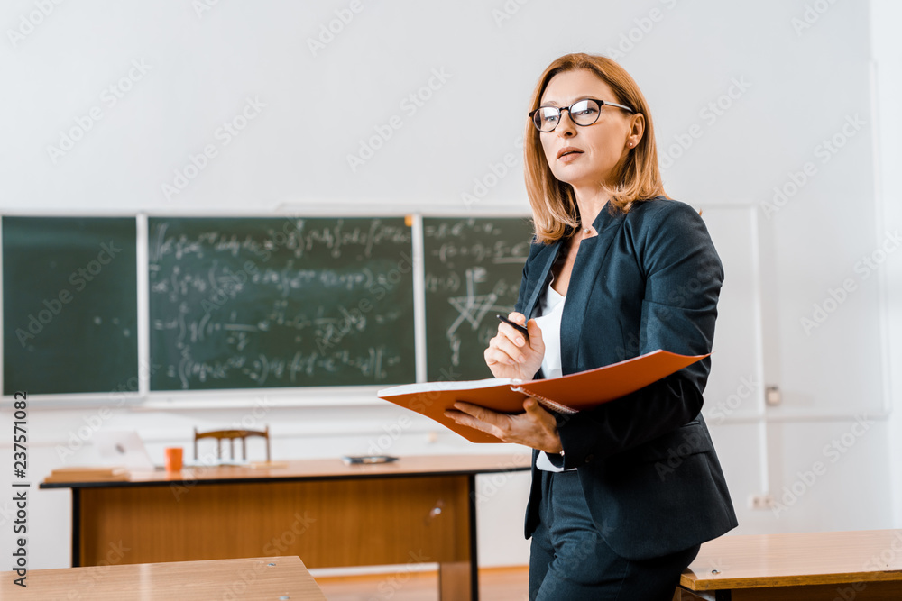 beautiful female teacher in formal wear and glasses holding notebook in classroom - obrazy, fototapety, plakaty 