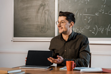 Wall Mural - smiling male teacher sitting at desk and showing laptop with blank screen in classroom