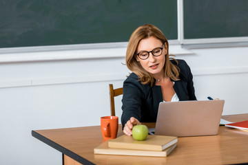 Wall Mural - beautiful female teacher in glasses sitting at computer desk and reaching for apple in classroom