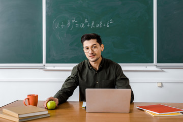 Wall Mural - male teacher in glasses sitting at computer desk, holding apple and looking at camera in classroom