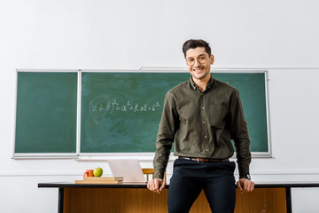 Wall Mural - smiling male teacher in formal wear looking at camera and standing near desk in classroom