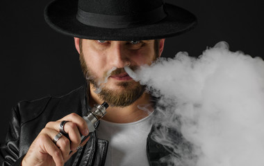 Man with vaping mod exhaling steam at black studio background. Bearded guy smoking e-cigarette to quit tobacco. Vapor and alternative nicotine free smoking concept