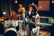 Mixed Race Woman Singing And Playing Guitar While Sitting On Chair With Legs Crossed. In Background Drummer, Saxophonist And Bass Guitarist.