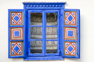 A traditional blue window on the white wall of a house in Dobrogea, Romania. Red and white geometrical motifs on the window shutters. 