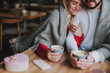 Cropped portrait of charming girl with closed eyes holding hand of her boyfriend while snuggling up to him. They sitting at the table and holding cups of cappuccino