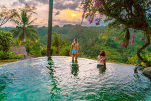 Two Women In Bathing Suits Sit On The Edge Of A Blue Tropical Pool Overlooking The Jungle