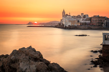 This Is A Sunset View With Rocks On The Foreground, The Mediterranean Sea Blurried By A Slow Shutter Speed And The Town Of Sitges In The Background.