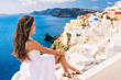 Europe summer travel destination Santorini tourist woman on vacation relaxing. Asian girl in white dress looking at famous white village Oia with the mediterranean sea and blue domes.