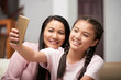 © DragonImages - Asian teenage girl with beautiful adult mother sitting on couch at home and smiling while taking selfie on phone