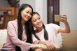 © DragonImages - Beautiful Asian adult woman with charming teen girl sitting on sofa and taking selfie with phone