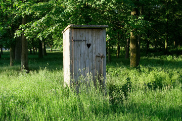 Old fashioned toilet building with wooden door standing outside among the grass with trees in the background.