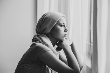 Wall Mural - Black and white photo of thoughtful young girl suffering from ovarian cancer wearing headscarf and looking through the window in medical center