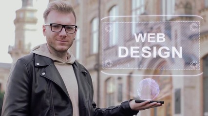 Smart young man with glasses shows a conceptual hologram Web Design. Student in casual clothes with future technology mobile screen on university background