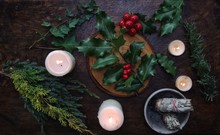 Yule Winter Solstice (Christmas) Themed Flat Lay With Branch Of Holly Plant On A Dark Wooden Table. Sage Smudge Sticks, White Lit Burning Candles, Dried Evergreens, Ivy Branches, Berries In Background