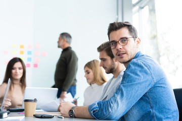 Wall Mural - Enjoying his work. Handsome young man looking at camera and smiling while his colleagues working in the background.