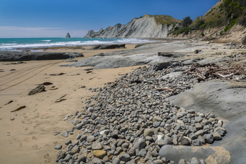 Low tide exposes a mix of small stones and solid rock above the sand beach and below towering cliffs along the sea in New Zealand 