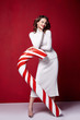 © indiraswork - Sexy pretty lady fashion model beautiful woman wear style skinny knitted white color dress celebration happy holidays merry Christmas Eve new year party hold candy cane caramel brunette makeup.