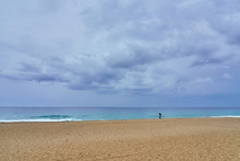 A Woman Under Umbrella On The Sandy Beach Looking At The Water Under Cloudy Sky On A Rainy Day.         