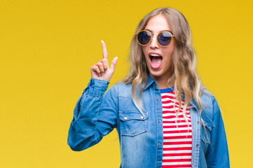 Wall Mural - Beautiful young blonde woman wearing sunglasses over isolated background pointing finger up with successful idea. Exited and happy. Number one.